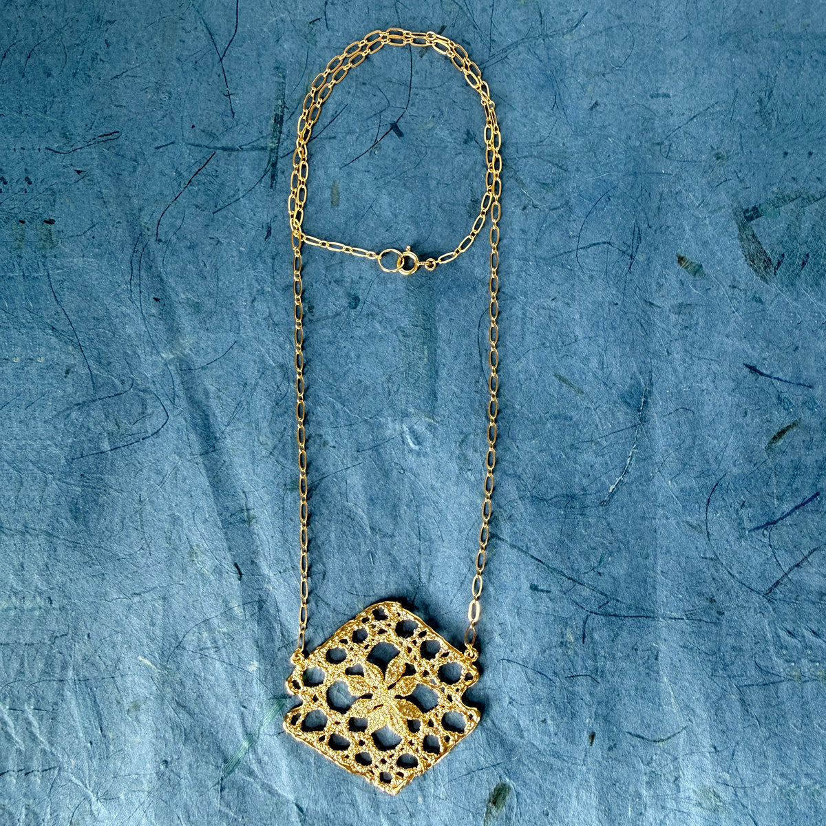 Lace pendant necklace in 24k gold on blue background.
