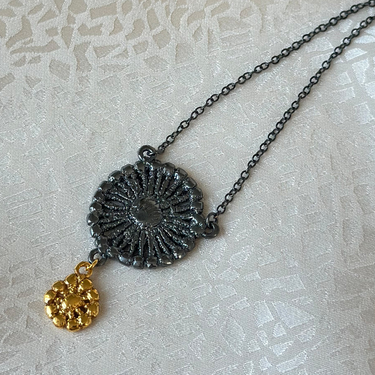 Necklace with black oxidized flower silver pendant and 24k gold charm on fabric background.