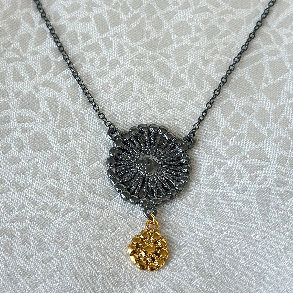 Necklace with black oxidized flower silver pendant and 24k gold charm on fabric background.